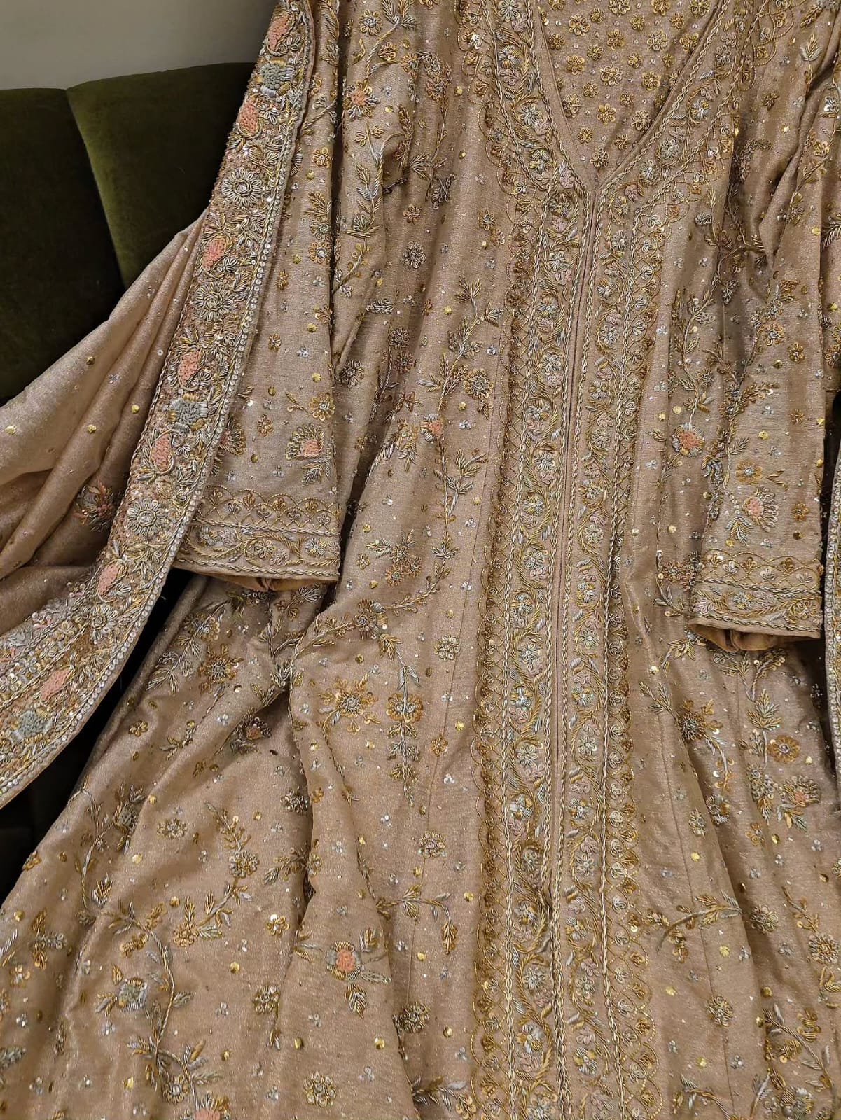 A close-up of an ornate beige or taupe traditional garment with extensive gold, silver, and pastel embroidered detailing, draped to show the fabric texture and pattern.