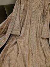A close-up of an ornate beige or taupe traditional garment with extensive gold, silver, and pastel embroidered detailing, draped to show the fabric texture and pattern.