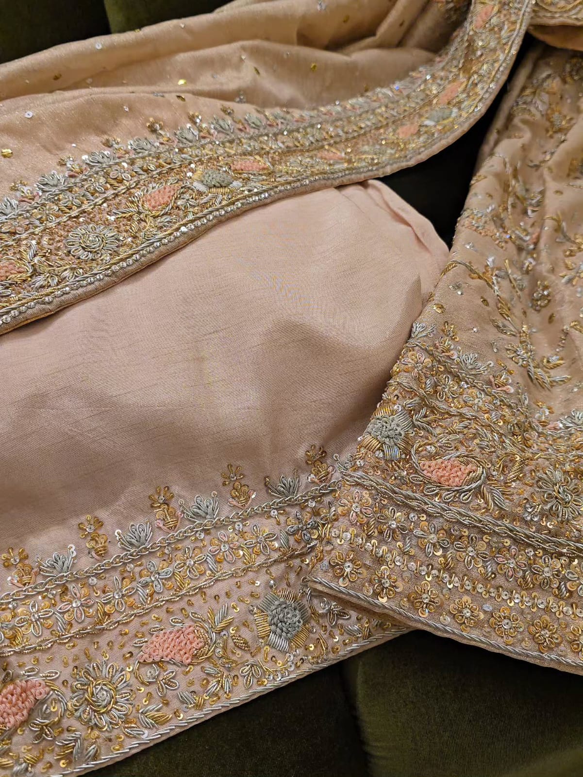 A close-up of a peach-colored traditional embroidered fabric set, featuring a solid fabric section and a heavily embellished border with metallic gold, silver, and pink floral embroidery.