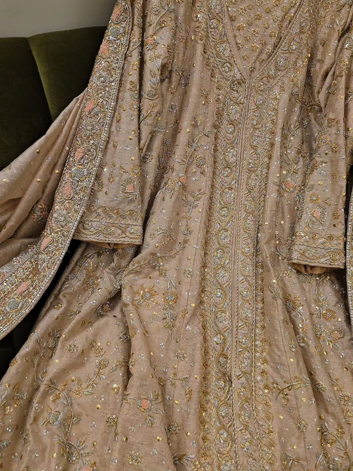 A close-up of an ornate beige or taupe traditional garment with extensive gold, silver, and pastel embroidered detailing, draped to show the fabric texture and pattern.