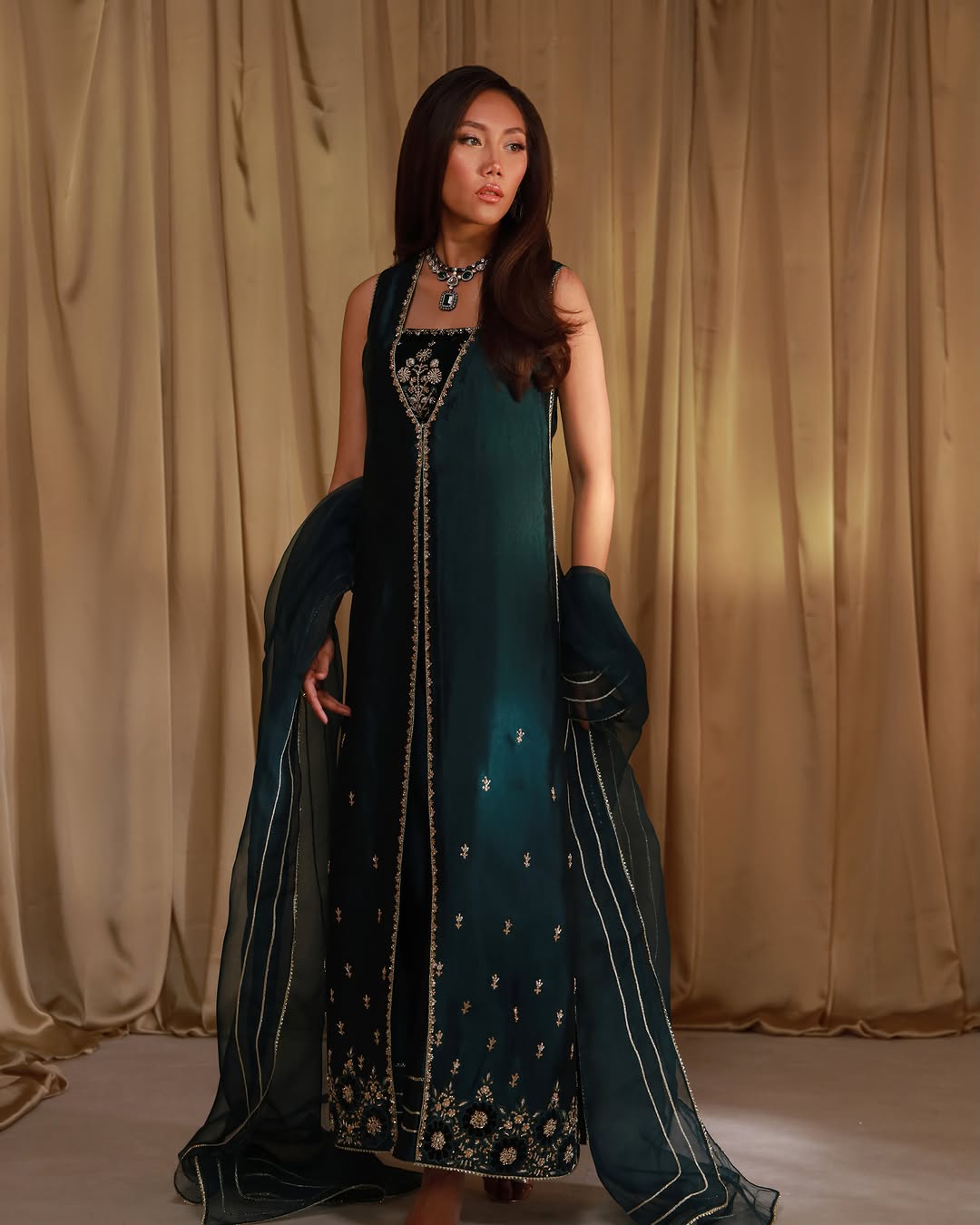 A woman models a dark teal green sleeveless Anarkali gown with intricate gold embroidery and a matching sheer dupatta, set against a draped beige fabric backdrop.
